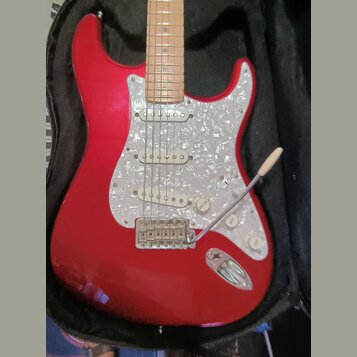 Červená elektrická kytara typu Stratocaster s bílo-perleťovým pickguardem v černém obalu. Na kytaru jsou instalovány tři snímače s bílými kryty a tři…