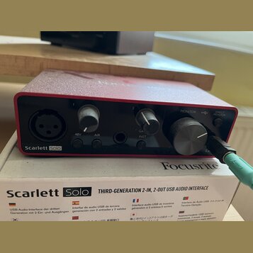 Červená USB audio rozhraní s nápisy Scarlett Solo a Focusrite. Na předním panelu jsou dva ovratové knofíky, jeden velký kruhový knofík MONITOR a vstu…