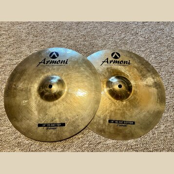 Dvě mosazné činely s viditelným rýhováním povrchu. Oba mají v horní části logo a nápis Armoni HANDMADE TURKISH CYMBALS. Levý činel má dole černý štít…