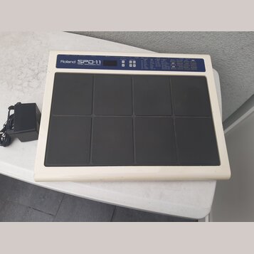 Bílá elektronická bicí pad s osmi černými polstrovanými plochami. V horní části je nápis Roland SPD-11 TOTAL PERCUSSION PAD, malý displej a ovládací…