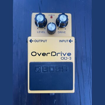 Žlutý efekt pedál s nápisem OverDrive OD-3. V horní části jsou tři ovládací knoflíky s popisky LEVEL a DRIVE a červená kontrolka CHECK. Ve spodní čás…