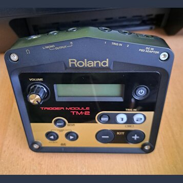 Černý trigger modul značky Roland, model TM-2. Zařízení má v horní části konektory s nápisy L/MONO OUTPUT, R OUTPUT, 1 TRIG IN, 2 TRIG IN a DC IN PSD…