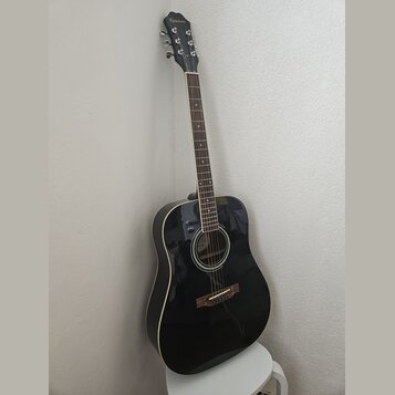 Černá akustická kytara s bílým lemováním korpusu a hnědým hmatníkem. Na hlavě je bílý nápis Epiphone. Nástroj je opřen o světlou stěnu a stojí na bíl…