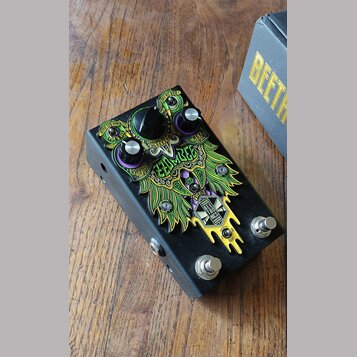 Černý efekt pedál s barevným grafickým motivem v zelené, žluté a fialové barvě. Na povrchu je nápis ZOOMBIE a logo s textem BEETLE JUICE PEDALS. Zaří…