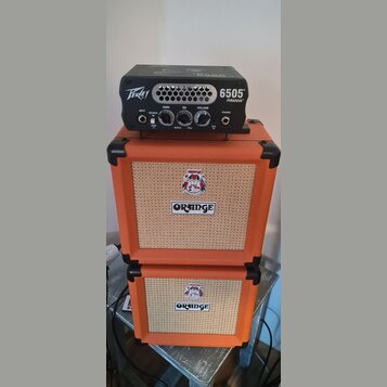 Černý kytarový zesilovač s nápisy Peavey a 6505 PIRANHA™ stojí na dvou oranžových reproduktorových boxech značky ORANGE. Zesilovač má ovládací prvky…
