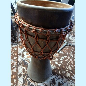 Djembe s tmavým dřevěným korpusem a napnutou kůží. Nástroj je omotaný hnědým provázkem v síťovém vzoru. Povrch dřeva vykazuje známky opotřebení a zap…