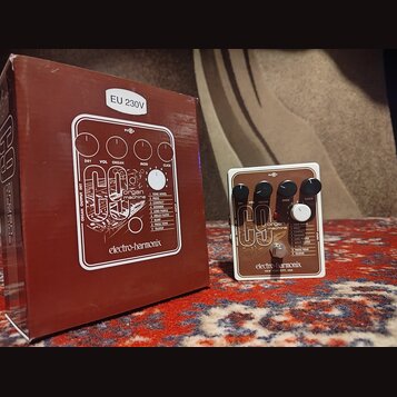 Na fotografii je efekt pedál Electro-Harmonix C9 Organ Machine v bílém rámu s hnědým předním panelem a čtyři černé ovládací knoflíky. Na pedálu jsou…