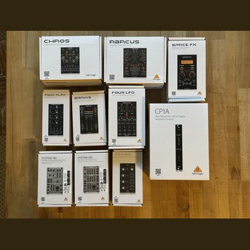 Fotografie zobrazuje deset bílých krabic s moduly pro Eurorack od značky behringer. Na krabicích jsou uvedeny názvy modelů: CHAOS, ABACUS, SPACE FX,…
