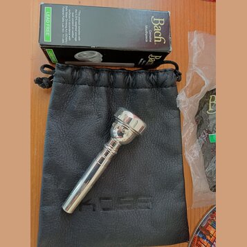 Stříbrný moutur z kovu položený na černém koženém vak s vyraženým nápisem KOSS. Nad nimi je černá krabička s bílými nápisy Bach Genuine Mouthpiece a…