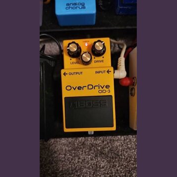 Žlutý efekt pedál s nápisy OverDrive a OD-3. Na horní části jsou tři černé ovládací knoflíky s nápisy LEVEL, TONE a DRIVE, nad nimi svítí červená kon…