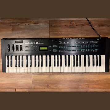 Černý digitální syntetizátor s bílými a černými klávesami. Na horním panelu je nápis YAMAHA DX11 a malý zelený displej s textem PLAY-SINGLE 004: Dim…