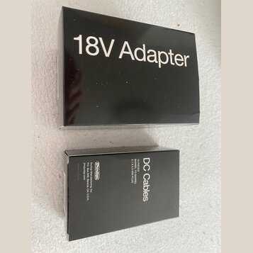 Černá krabička s bílým nápisem 18V Adapter. Vedle ní je menší černá krabička s bílým nápisem DC Cables a textem Dunlop Manufacturing, Inc. P.O. Box 6…