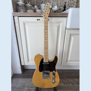 Elektrická kytara s přírodním dřevěným povrchem těla a hmatníkem. Na hlavě je viditelné logo Fender. Kytara má černý pickguard a chromované potenciom…