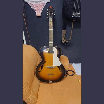 Polodupatá elektrická kytara s korpusem v přechodové barvě od žluté po černou. Nástroj má bílý plechový kryt (pickguard) a bílé ladičky na hlavě. Na…