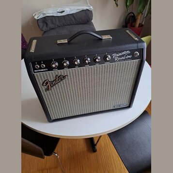 Černý kytarový kombozesilovač s šedou textilní maskou reproduktoru. Na horním ovládacím panelu jsou nápisy Princeton Reverb Amp, FENDER MUSICAL INSTR…