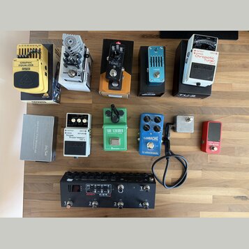 Sestava efektů pro kytaru rozložená na dřevěném povrchu. V horní řadě jsou pedály Behringer GRAPHIC EQUALIZER EQ700, AMPLITUBE, TONEX, S-BAND a BOSS…