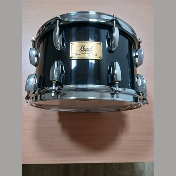 Černý buben s chromovaným obručováním a ladidly. Na korpusu je zlatý štítek s nápisy Pearl, MAPLE SHELL, 000170 a MADE IN JAPAN. Povrch nástroje je g…