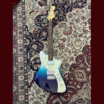 Elektrická kytara s trupem v modrém přechodovém odstínu (fade) od tmavě modré po světle modrou. Na hlavě hmatníku je viditelné logo Fender. Nástroj m…