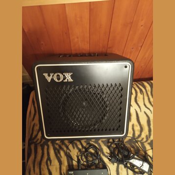 Černý kytarový kombo zesilovač s bílým rámečkem a mřížkou z kovového pletiva. Na přední straně je umístěno stříbrné logo VOX. Před přístrojem leží če…