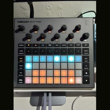 Šedý sampler Novation Circuit Rhythm s mřížkou podsvícených padů v modrých a oranžových barvách. V horní části jsou umístěny ovládací knoflíky s popi…