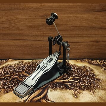 Černý bicí pedál s kovovým šlapadlem a stříbrnými detaily. Na šlapadle je viditelné logo MAPEX v oválném rámečku. Pedál vykazuje mírné známky používá…