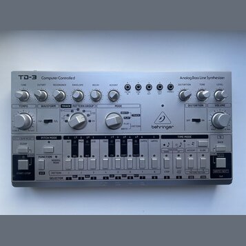 Stříbrný analogový basový syntetizátor s nápisy TD-3 Computer Controlled a Analog Bass Line Synthesizer. Na pravé straně je umístěno logo a značka be…