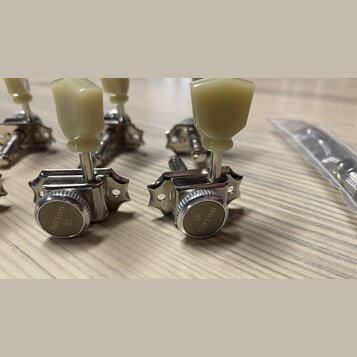 Detailní pohled na čtyři chromované ladicí mechanismy s krémovými plastovými knoflíky. Na kovových krytech je vyryt nápis GOTOH a logo ve tvaru styli…