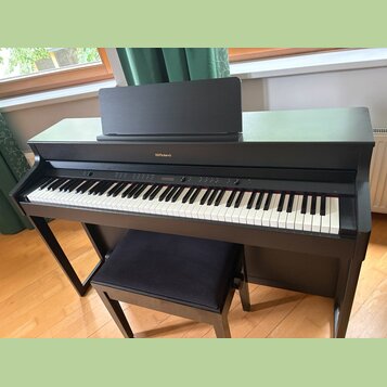 Černý digitální piano v podobě konzole s přiloženým polstrovaným černým stolečkem. Na přední horní hraně je umístěn nápis Roland. Nástroj má standard…