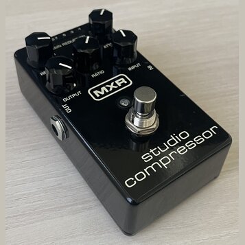 Černý efekt pedál s nápisy MXR a studio compressor. Na horní straně se nachází pět černých otočných ovladačů s bílými značkami a popisky RE, RATIO, A…