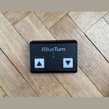 Černý obdélníkový ovladač s nápisem iRig BlueTurn. Zařízení má dvě bílá čtvercová tlačítka s černými šipkami směřujícími nahoru a dolů, nad nimi jsou…