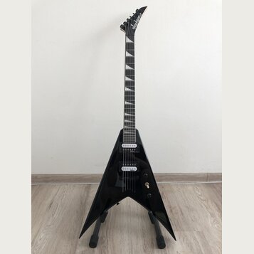 Černá elektrická kytara s tvarem V umístěná na stojanu. Na hlavě hmatníku je bílý nápis Jackson. Kytara má dva humbuckery, ladicí kolíky na hlavě a b…