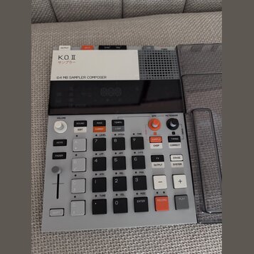 Šedý sampler s černým displejem a ovládacími prvky. Na horní bílé části je nápis K.O. II, pod ním japonské znaky サンプラー a text 64 MB SAMPLER COMPOSER.…