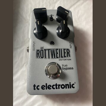 Stříbrný efekt pedál s nápisy RÖTTWEILER DISTORTION, true bypass a tc electronic®. Na horní části jsou čtyři černé oválné ovládací knoflíky a nápisy…