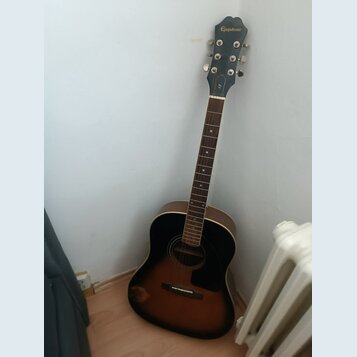 Akustická kytara s tmavým korpusem v provedení sunburst. Na hlavě hmatníku je bílý nápis Epiphone. Nástroj je opřen o bílou zeď vedle radiátoru.