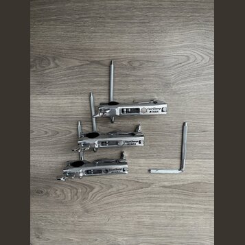 Tři kovové stříbrné svorky s černými gumovými vložkami. Každá svorka má bílý štítek s nápisem FastClamp TAMA. Povrch kovových částí je lesklý a v dob…