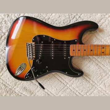 Elektrická kytara s trupem v barevnosti sunburst a černým pickguardem. Na kytare jsou tři snímače typu single-coil, chromovaný most s páčkou a tři ov…