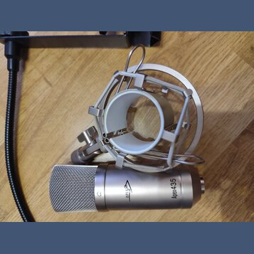 Stříbrný kondenzátorový mikrofon s mřížkou a doprovodným šokovým držákem z kovu. Na těle mikrofonu je nápis Apex4.35 a logo APEX. Povrch nástrojů je…