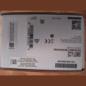 Fotografie znázorňuje bílou papírovou krabici s černými nápisy a logy. V pravém horním rohu je logo SHURE a nápis Made in China. Na krabici jsou uved…