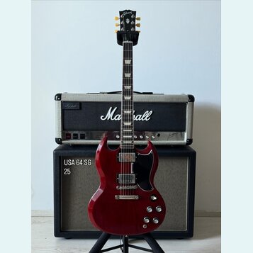 Elektrická kytara v tmavě červené barvě s černým pickguardem, umístěná na stojanu. Na hlavě hmatníku je nápis Gibson a na kombu v pozadí značka Marsh…