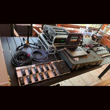 Na černém stole je rozložena sada hudebního vybavení. V popředí je stříbrný MIDI kontrolér s nápisem FCB1010 MIDI FOOT CONTROLLER a řadou černých spí…