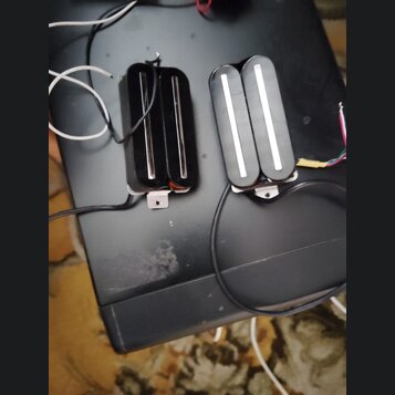 Dvě černé humbucker snímače s bílými plastovými krytkami nad pólky. Snímače jsou vybaveny připojovacími kabely a kovovými montážnímideskami. Na povrc…