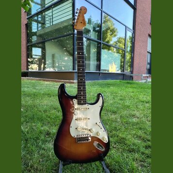 Elektrická kytara s tmelem v barvě sunburst, bílým pickguardem a hnedým hmatníkem. Na hlavě je viditelné logo Fender. Nástroj má tři snímače, pětipos…