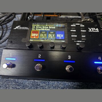 Černý kytarový procesor s nápisy FRACTAL AUDIO SYSTEMS a VP4 VIRTUAL PEDALBOARD. Zařízení má digitální displej s texty UI: Drive Drive Drive!, S1: Ki…