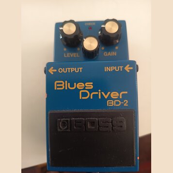 Modrý efekt pedál s nápisy Blues Driver a BD-2. Na spodním černém spínači je vyražené logo BOSS. Horní část obsahuje tři ovládací knoflíky s popisky…