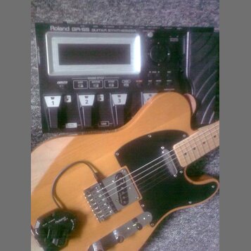 Černý kytarový syntetizátor s nápisem Roland GR-55 GUITAR SYNTHESIZER a displejem. Před ním leží kytara s přírodním dřevěným povrchem, černým pickgua…