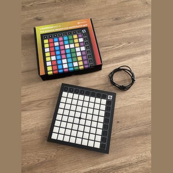 Na fotografii je černý MIDI kontrolér Novation Launchpad X s mřížkou bílých podsvícených polí a ovládacími tlačítky po stranách. Vedle zařízení leží…