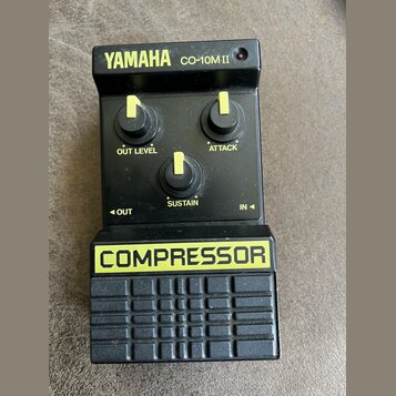 Černý kompresor s nápisem YAMAHA CO-10M.II v horní části a žlutým nápisem COMPRESSOR na spodním panelu. Zařízení má tři černé oválné knoflíky s žlutý…
