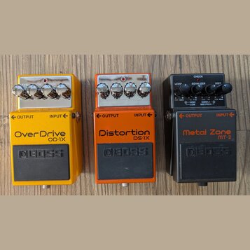 Tři kytarové efekty značky BOSS v žluté, oranžové a černé barvě. Horní pedál má nápisy OverDrive OP-1X, prostřední Distortion DS-1X a spodní Metal Zo…