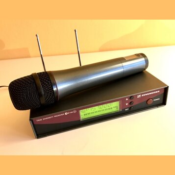 Bezdrátový mikrofon s černou mřížkou a stříbrno-šedým tělem leží na černém přijímači. Na přijímači je nápis TRUE DIVERSITY RECEIVER ew 100 G2, logo a…