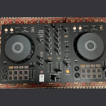 Černý DJ kontrolér s dvěma jog-koly a centrálním mixážním sekcí. Na přístroji jsou viditelné nápisy PERFORMANCE DJ CONTROLLER, DDJ-FLX4, rekordbox a…